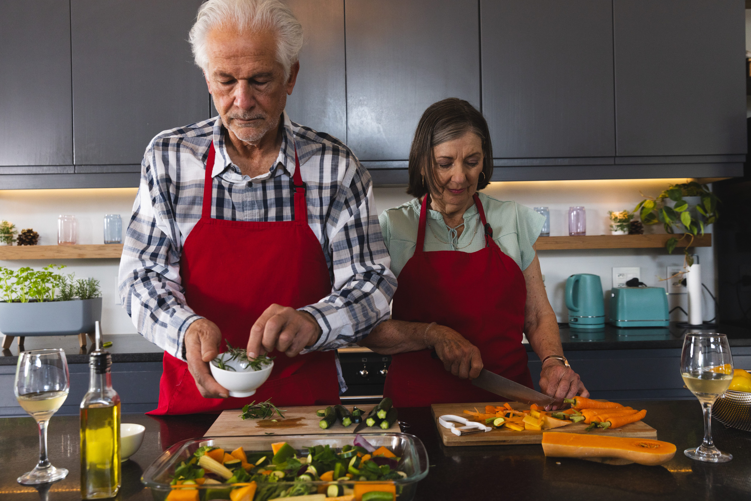 Senior couple cooking at kitchen island wearing red aprons, sprinkling herbs and slicing carrots. Cooking, kitchen, gourmet, culinary, preparation, rustic, wellness