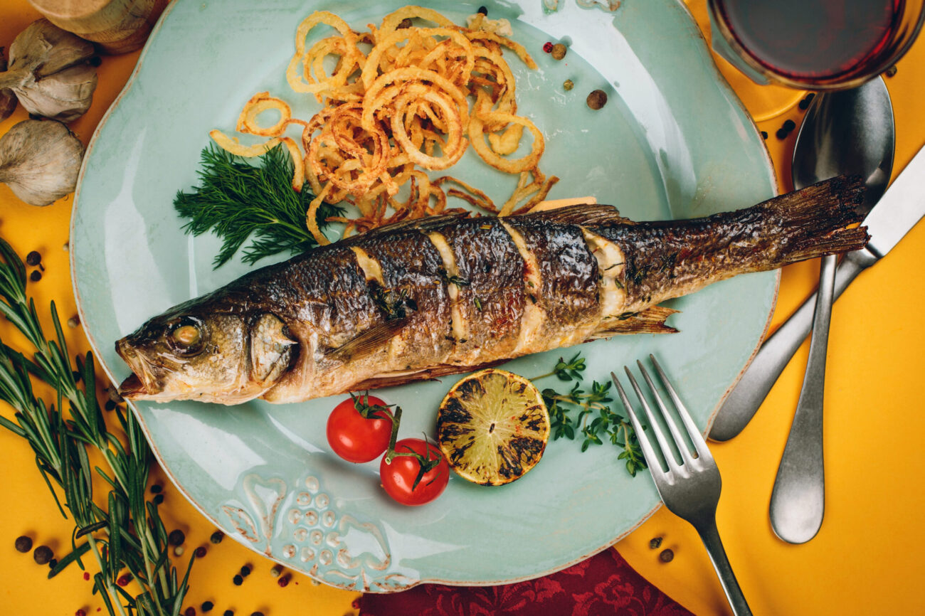A delectable plate showcasing a perfectly grilled fish, surrounded by crispy onion rings, fresh herbs, and vibrant cherry tomatoes, all set against a cheerful yellow background that invites a culinary adventure. Image Credit: kaninstudio/ 123RF