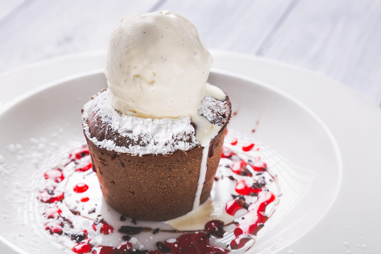 A decadent chocolate lava cake, warm and gooey, topped with a generous scoop of creamy vanilla ice cream, served on a plate with a drizzle of raspberry sauce, promising a delightful indulgence for dessert lovers. Image Credit: gar1984/ 123RF