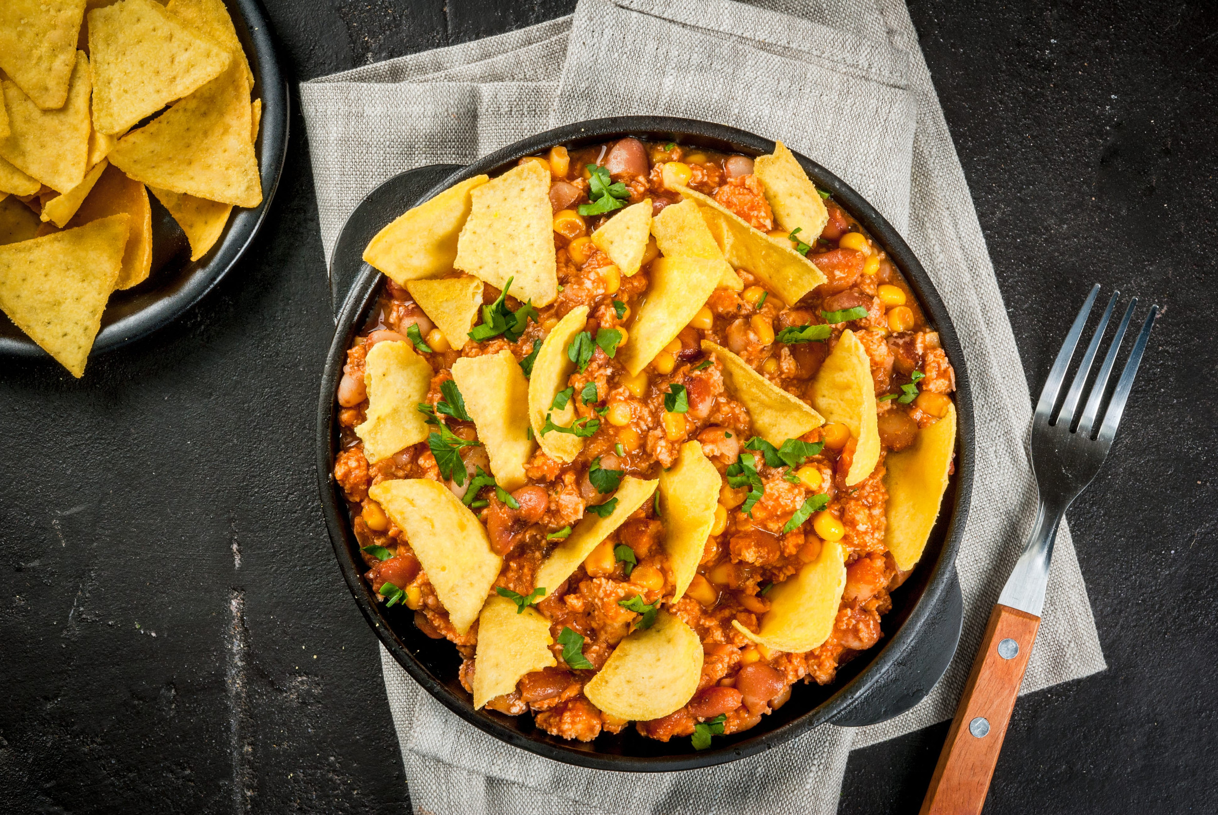 Traditional Mexican Chilean food, chili con carne, served in a portioned skillet pan with nachos tortilla. On black concrete table, top view copy space