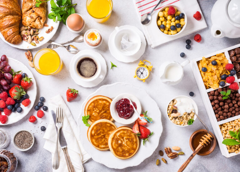 A delightful breakfast spread featuring golden pancakes topped with fresh strawberries and berry jam, surrounded by croissants, vibrant fruits, and refreshing beverages, perfect for starting the day with energy and flavor. Image Credit: imelnyk/ 123RF