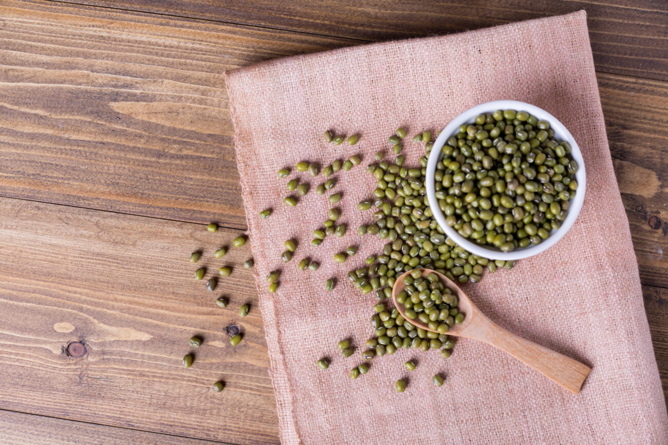 A rustic kitchen scene featuring fresh green mung beans spilling from a bowl onto a charming linen cloth, exuding a sense of natural abundance and culinary potential. Image Credit: iamjazzdog/ 123RF