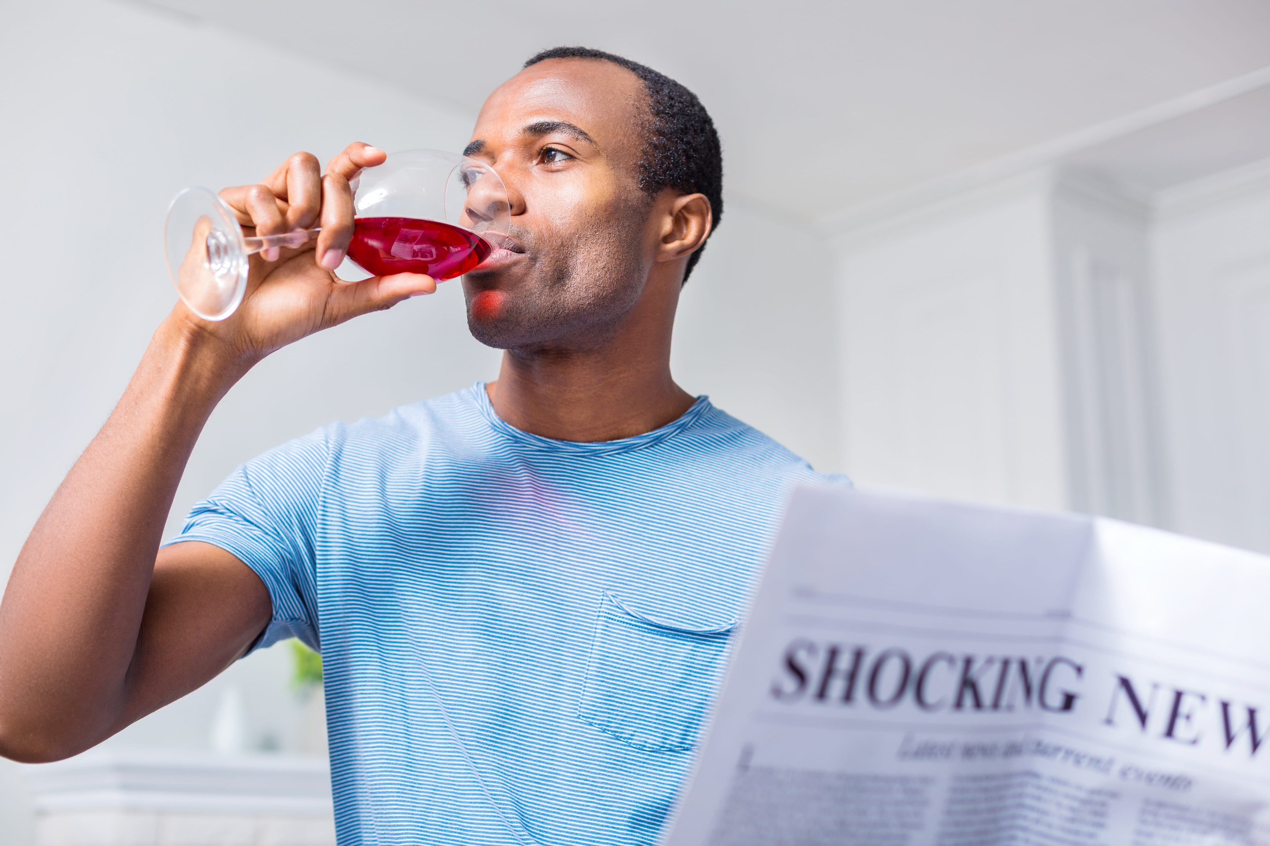 Alcohol addiction. Nice pleasant adult man holding a newspaper and sipping wine while drinking alcohol