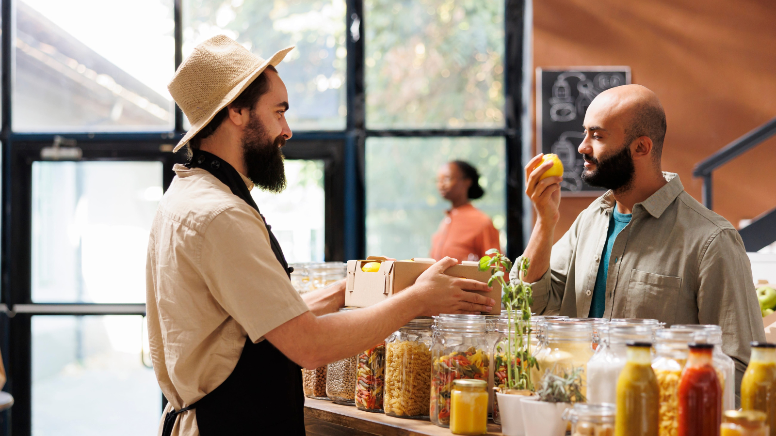 Middle Eastern male customer smells yellow fruit, that looks like a lemon, while caucasian storekeeper wearing a hat and apron looking over to the other side of counter holding a package. Image Credit: dragoscondrea/ 123RF