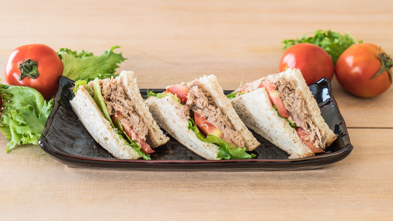 A delectable trio of freshly made sandwich slices, layered with tender tuna, crisp lettuce, and ripe tomatoes, served on a sleek black platter, surrounded by vibrant tomatoes and greens that add a touch of freshness to any meal setting. Image Credit: topntp/ 123RF