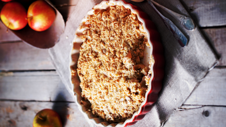 A warm apple crumble fresh from the oven, its golden crust invitingly crisp. Nestled in a rustic red dish, this delightful dessert is surrounded by freshly picked apples, evoking a sense of cozy autumn afternoons and sweet family gatherings. Image Credit: paviogel/ 123RF
