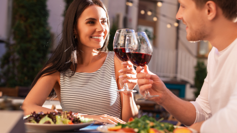 In a cozy, outdoor caf&eacute;, a couple enjoys a romantic dinner while toasting their glasses of red wine. The soft glow of string lights adds warmth to the evening, creating a perfect ambiance for love and laughter under the stars. Image Credit: yacobchuk/ 123RF