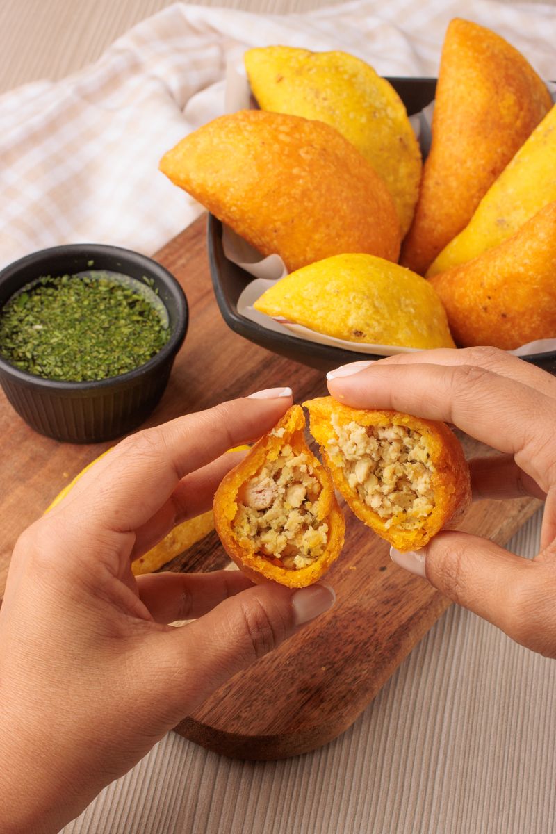 A person holds a Colombian empanada filled with beef and spices, alongside a dipping sauce.