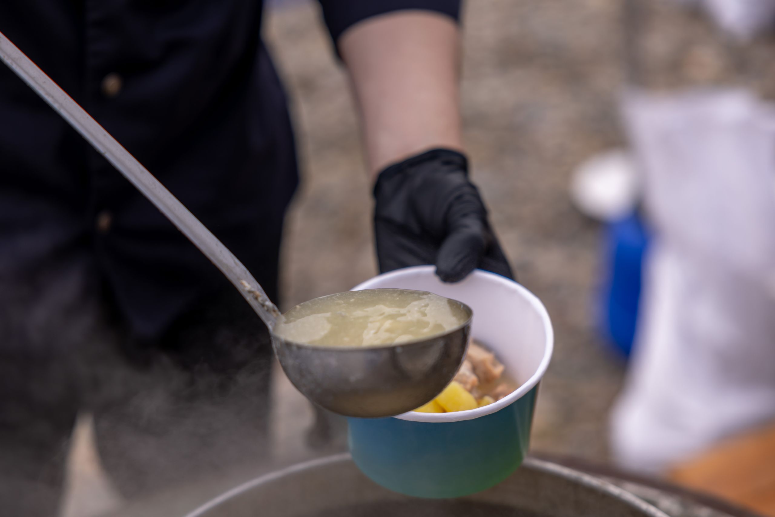 A person serves steaming hot soup from a pot into a bowl using a ladle outdoors.