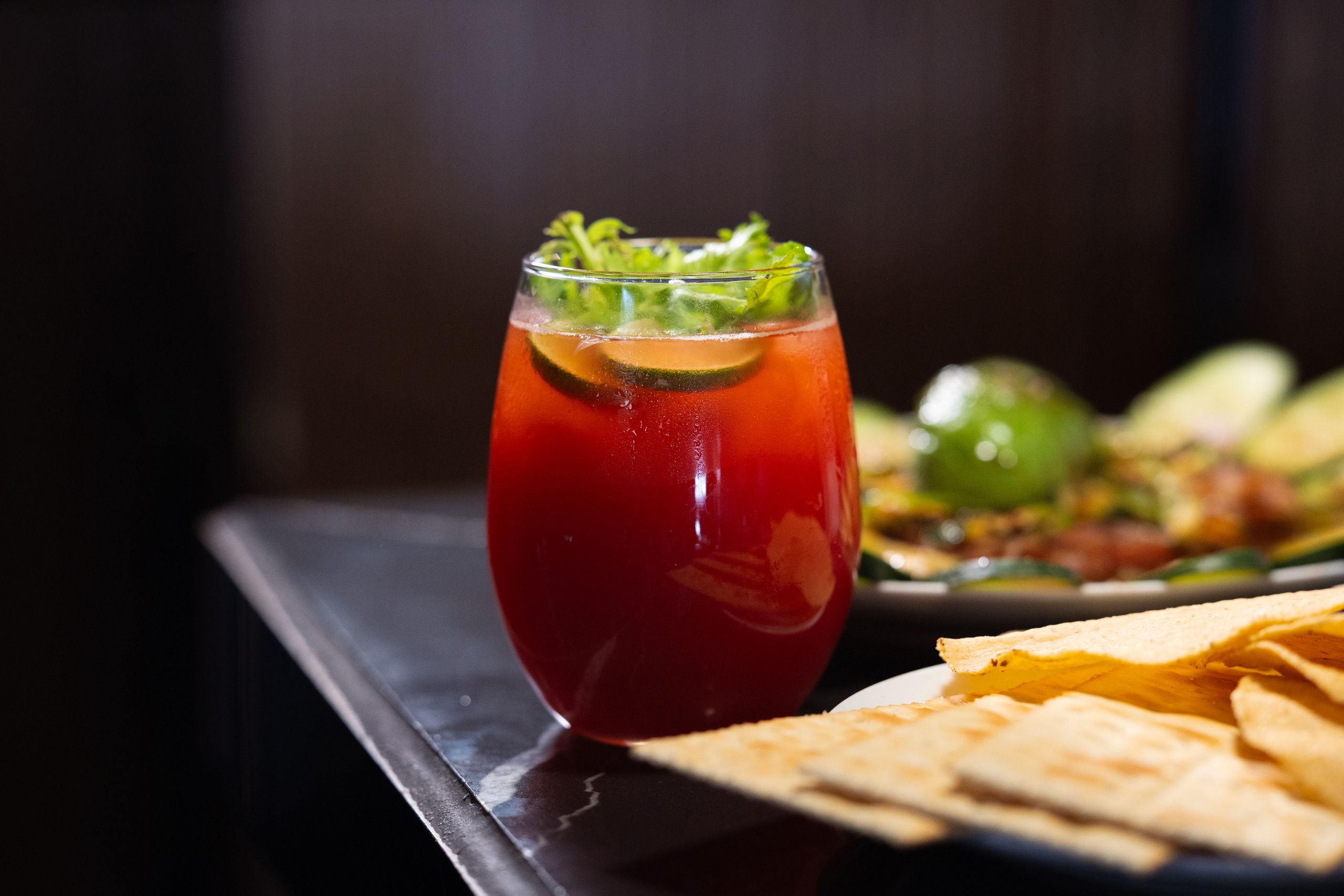 A vibrant red cocktail garnished with lime and cilantro, served with colorful appetizers.