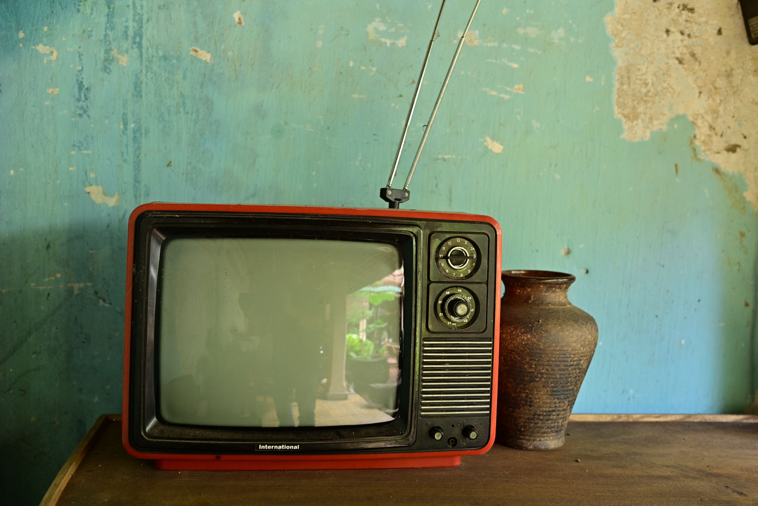 A vintage television with antenna beside a ceramic vase on a worn wooden surface against a rustic wall.
