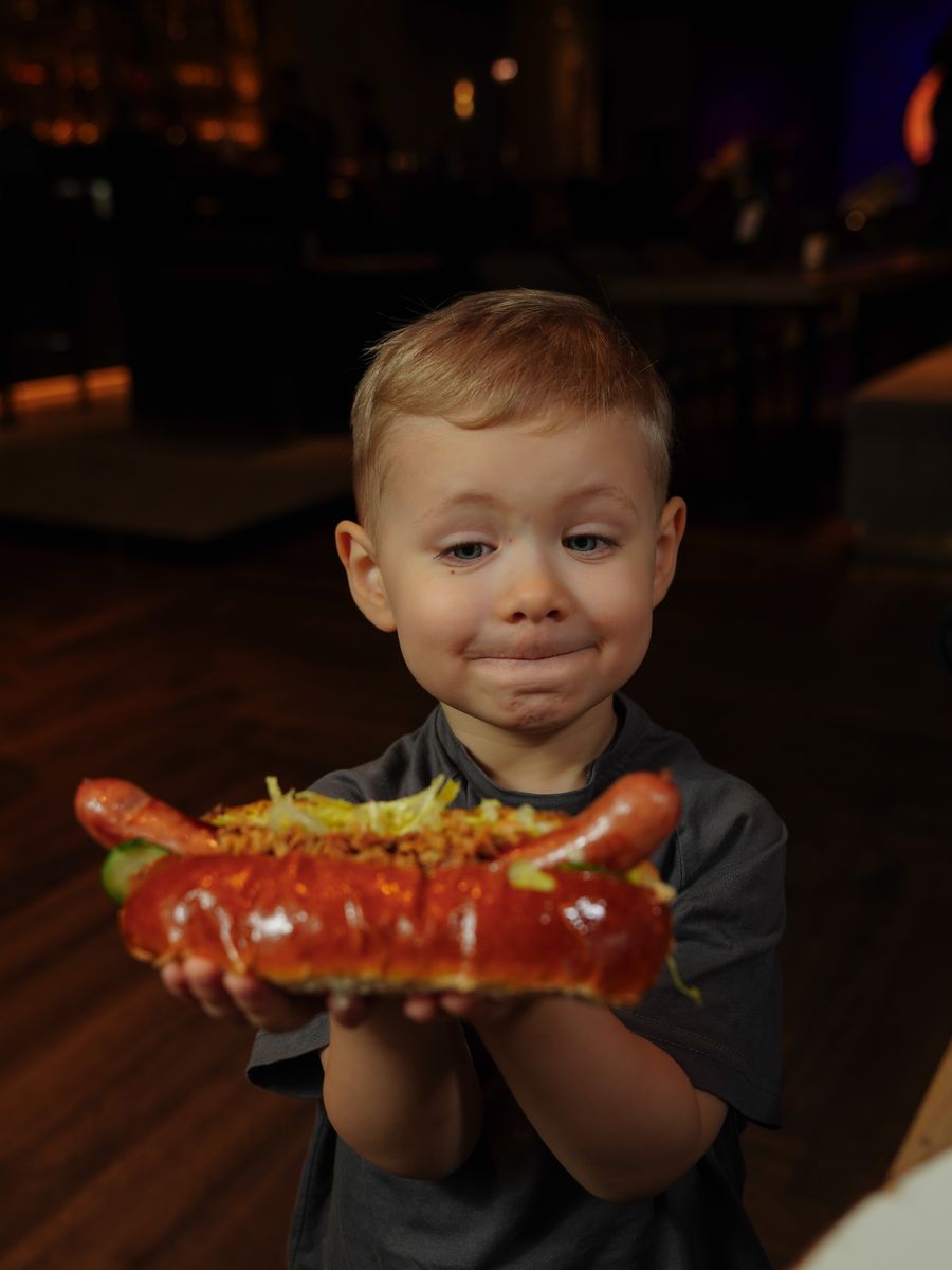 Adorable child proudly holds a large, delicious hot dog with toppings in a cozy restaurant setting.