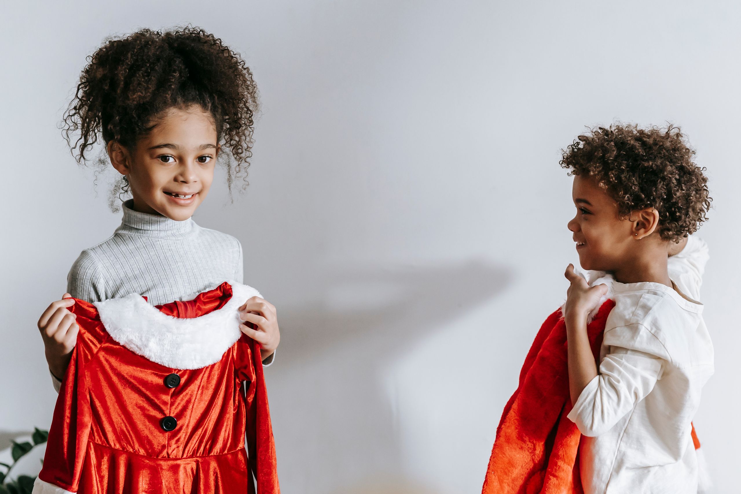Cheerful African American siblings with red Santa Claus outfits in hands standing in light room at home during Christmas holiday
