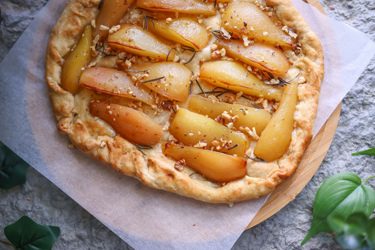 Close-up of a delicious homemade pear galette, topped with chopped nuts and rosemary, on a textured surface.