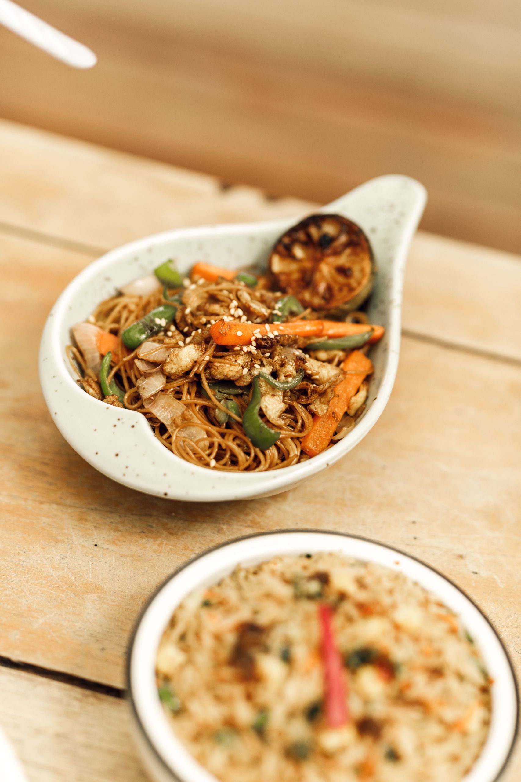 Close-up of an Asian noodle dish with fresh vegetables and grilled lemon, styled in a ceramic bowl.