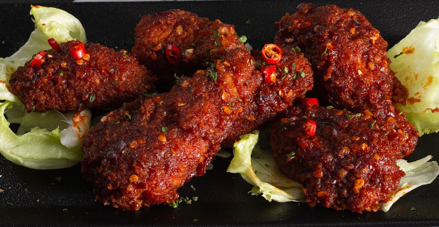 Close-up of spicy fried chicken wings on a dark plate, garnished with lettuce and herbs.