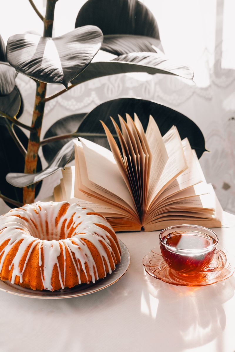 Cozy morning setup with a bundt cake, hot tea, and a book beside a ficus plant.