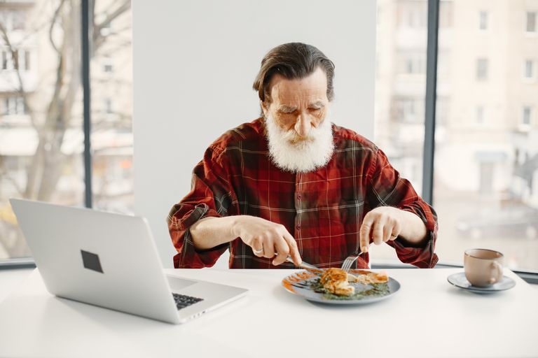 Elderly man in checkered shirt eating lunch at a table with a laptop and coffee indoors.