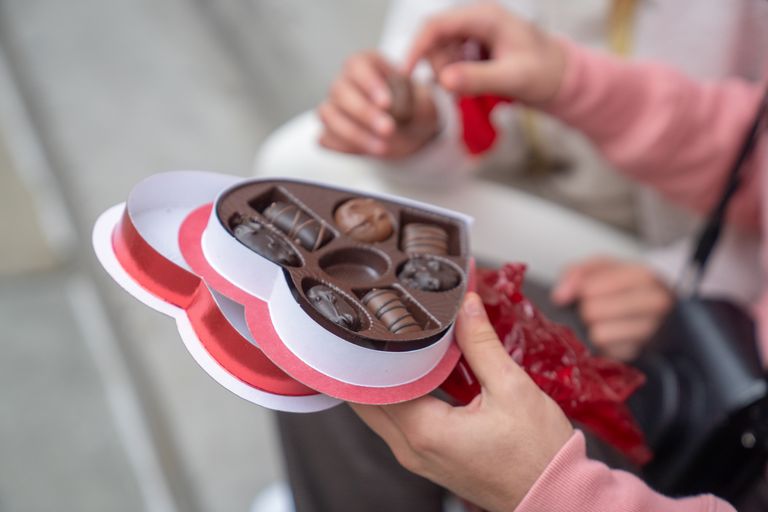 From above of crop unrecognizable young man holding heart shaped gift box and giving tasty chocolate to girlfriend while sitting together on street on Saint Valentines Day