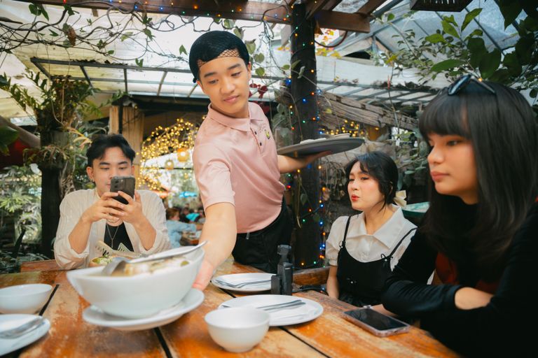 Group of friends being served by a waiter in an outdoor restaurant setting with festive lights.