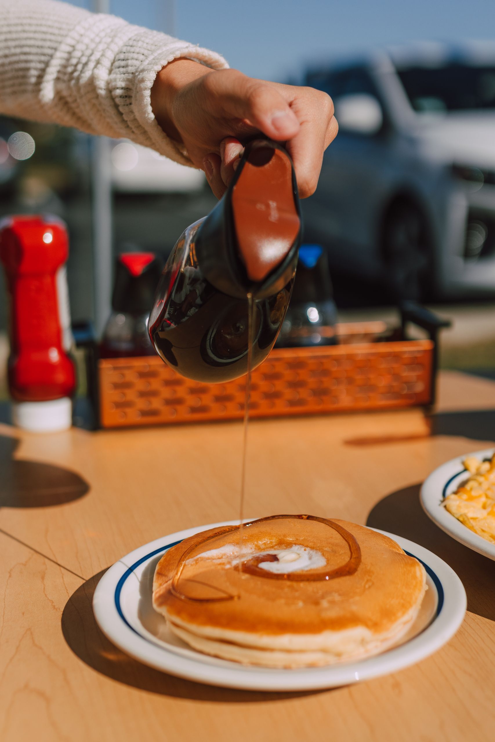 Hand pouring maple syrup onto pancakes outdoors on a sunny day.
