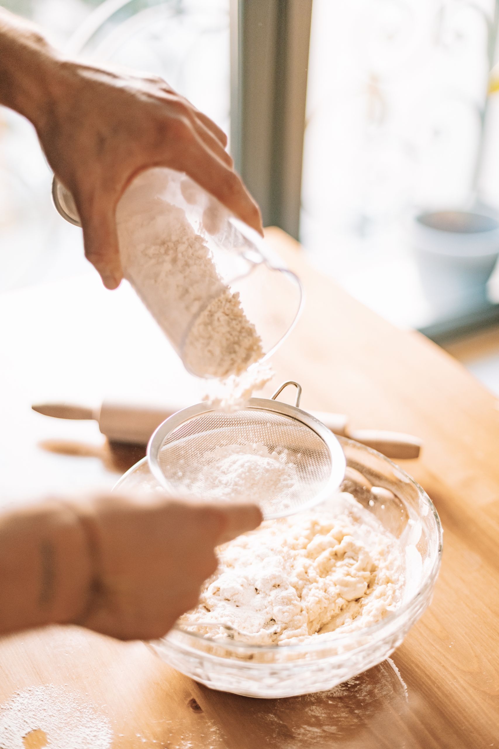 Hands sifting flour through a sieve over a glass bowl to prepare dough in a warm kitchen light.