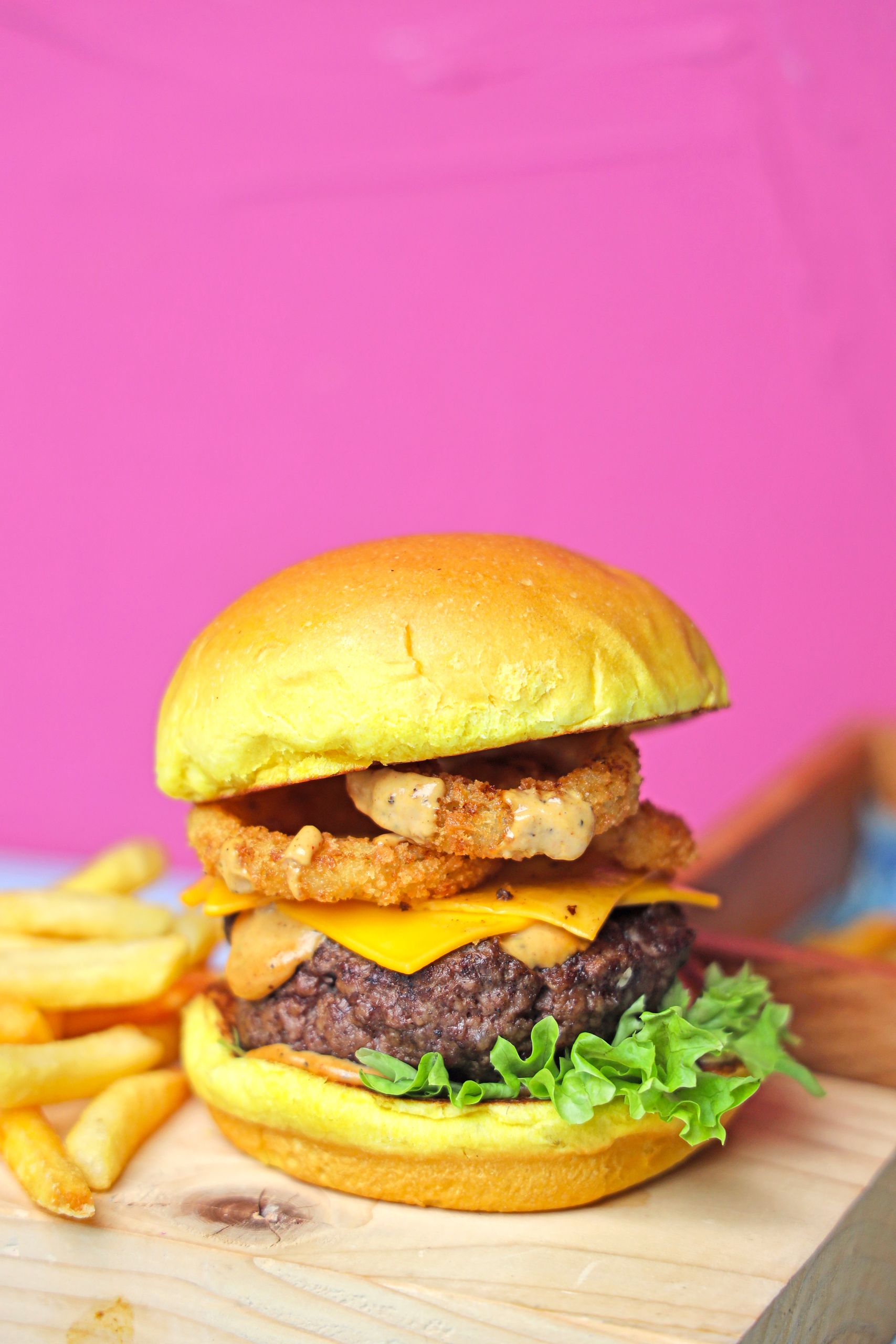 Juicy cheeseburger topped with onion rings on a wooden board with french fries.