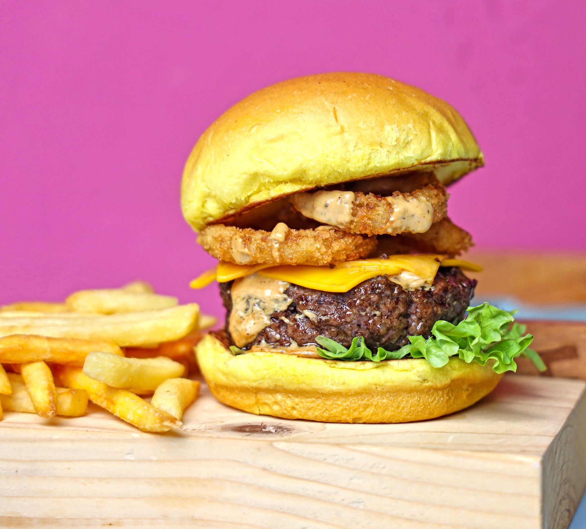 Juicy cheeseburger with onion rings and fries served on a pink background.