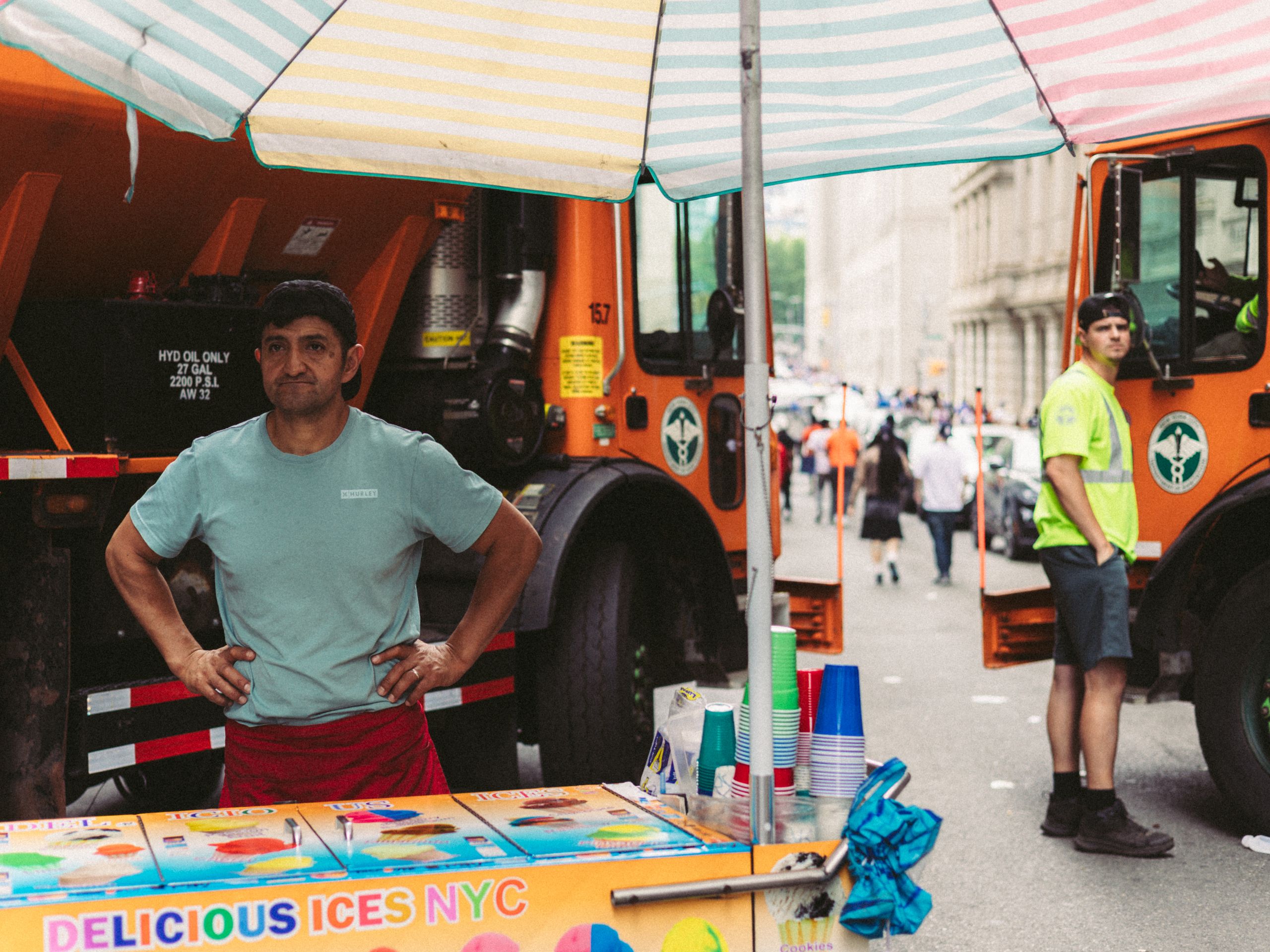 New York City street vendor selling ices beside construction trucks on a vibrant summer day.