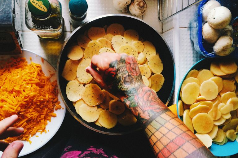 Overhead view of a tattooed arm preparing potato and cheese dish on a kitchen counter.