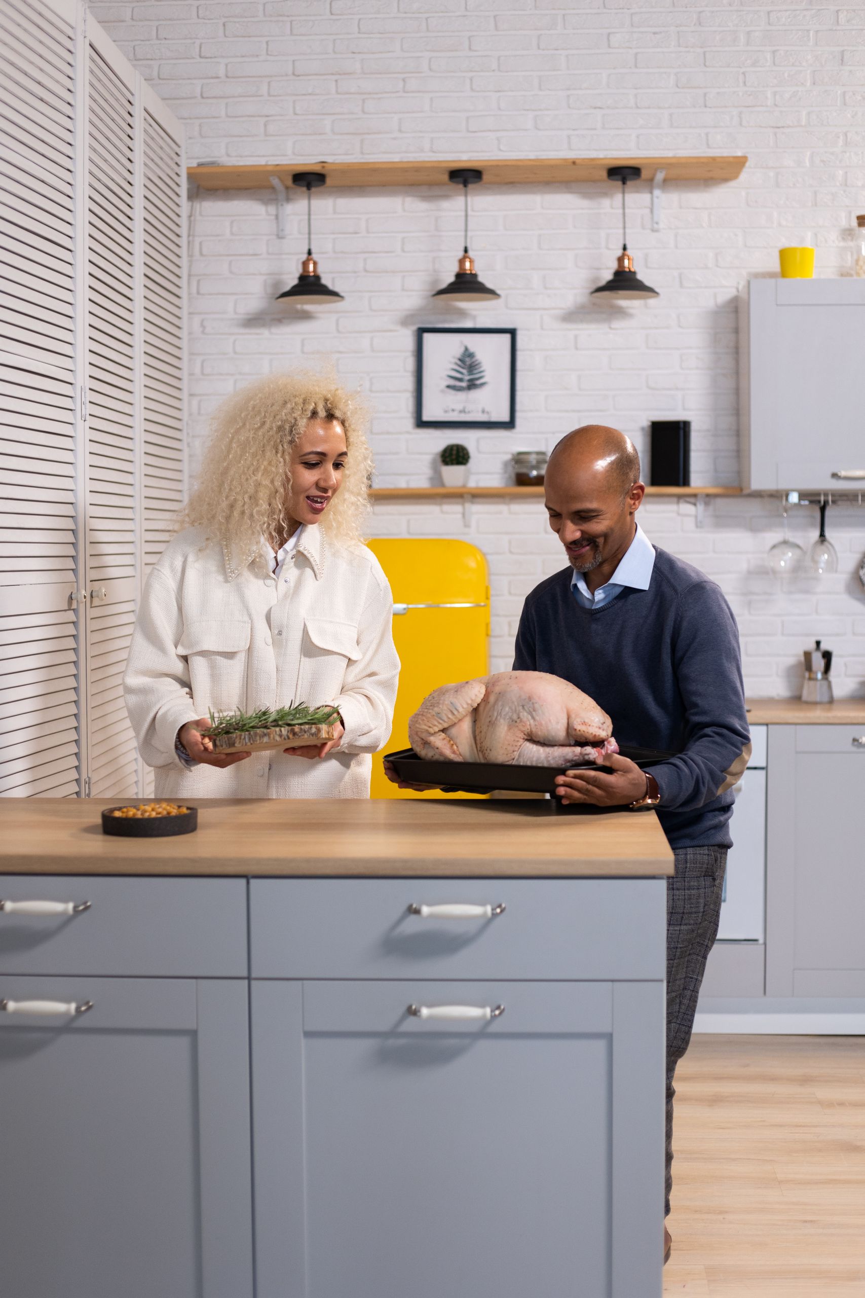 Positive African American wife with rosemary and husband with tray of whole raw turkey standing at counter in kitchen while cooking
