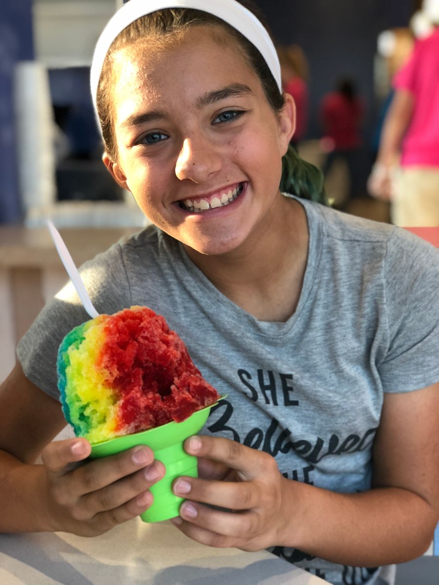 Smiling girl enjoys a vibrant rainbow snow cone indoors, radiating happiness and youth.