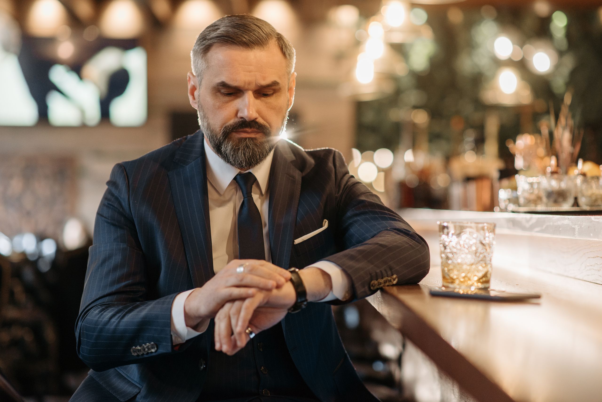 Stylish businessman in corporate attire enjoying whiskey at a bar, checking time.