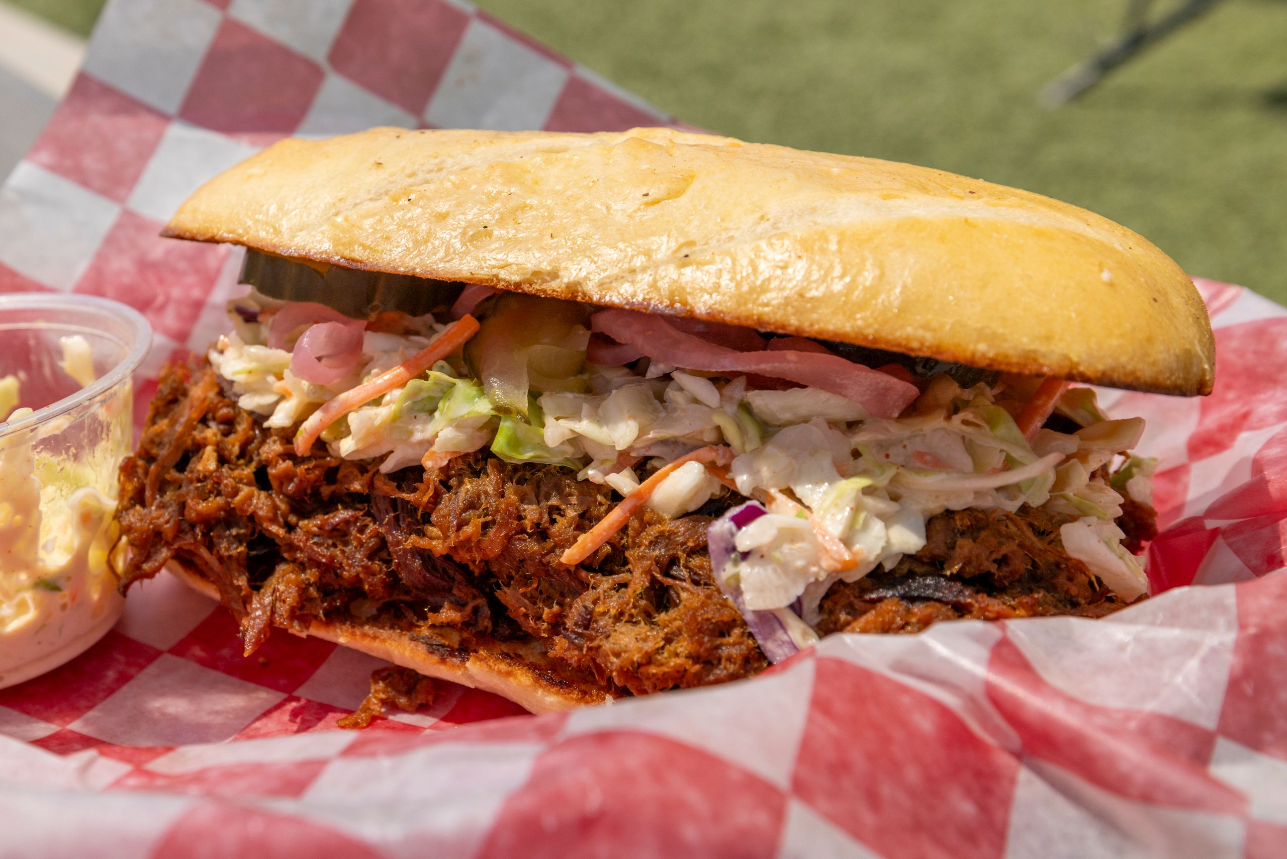 Tasty pulled pork sandwich topped with coleslaw on checkered paper outdoors.