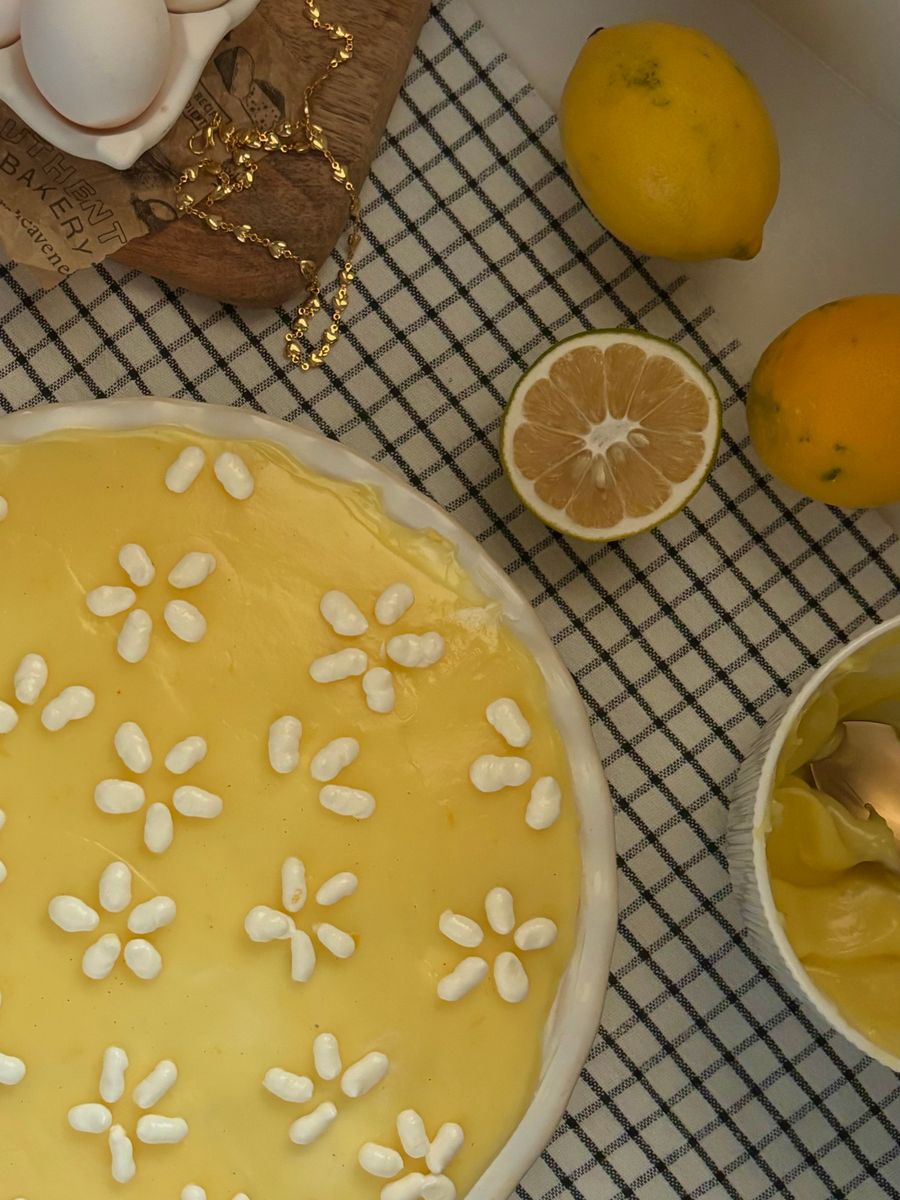Top-down view of a freshly baked lemon pie with decorative flower patterns, lemons, and baking setup.
