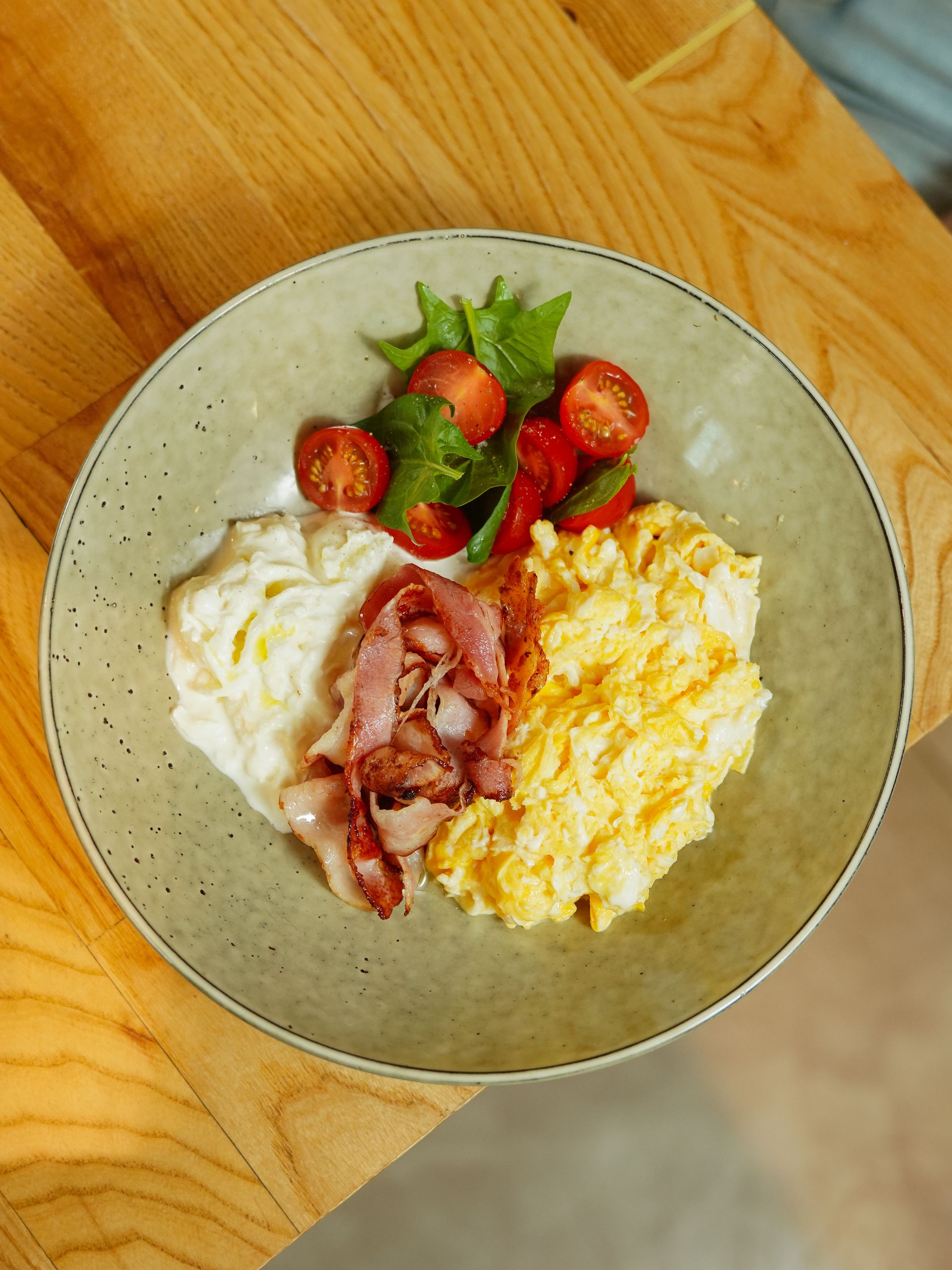 Top view of a delicious homemade breakfast featuring scrambled eggs, bacon, cherry tomatoes, and greens.
