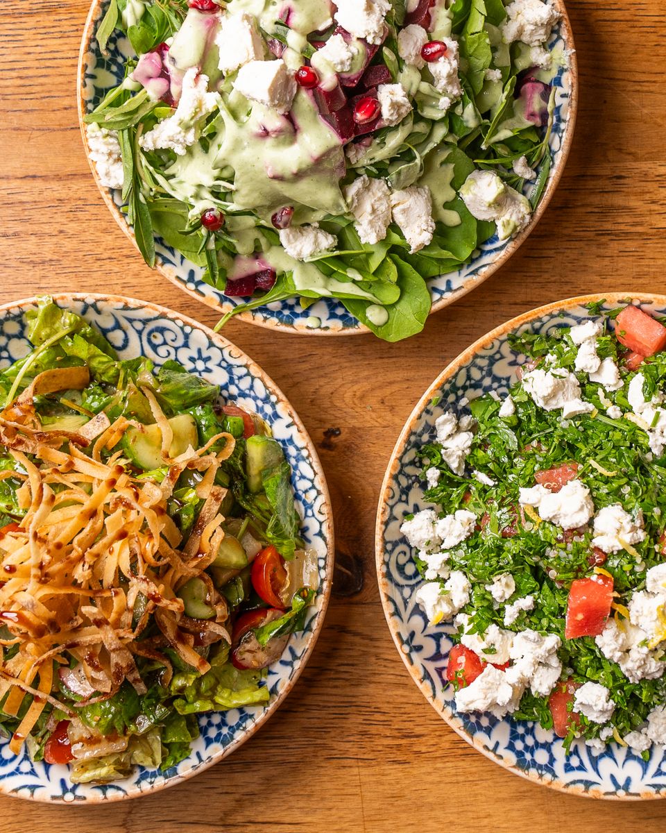 Top view of three fresh salads with feta cheese, vegetables, and herbs on a wooden table.