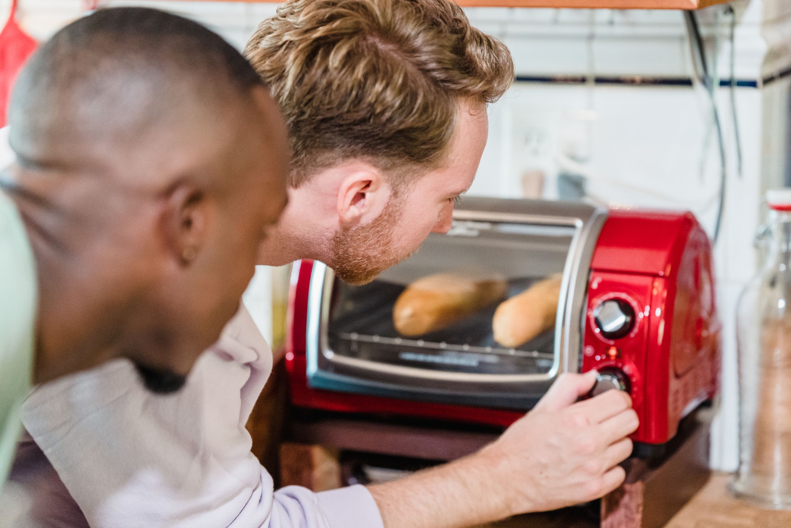 Two men closely monitor bread baking in a red toaster oven for breakfast.