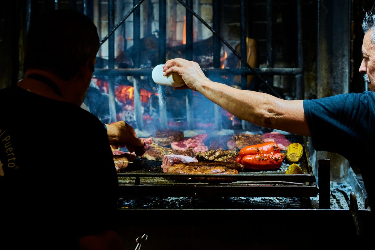 Uruguayan chefs grilling meats and vegetables over open flame in an indoor parrilla.